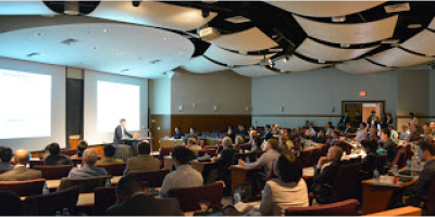 A speaker lectures to a crowded conference room.